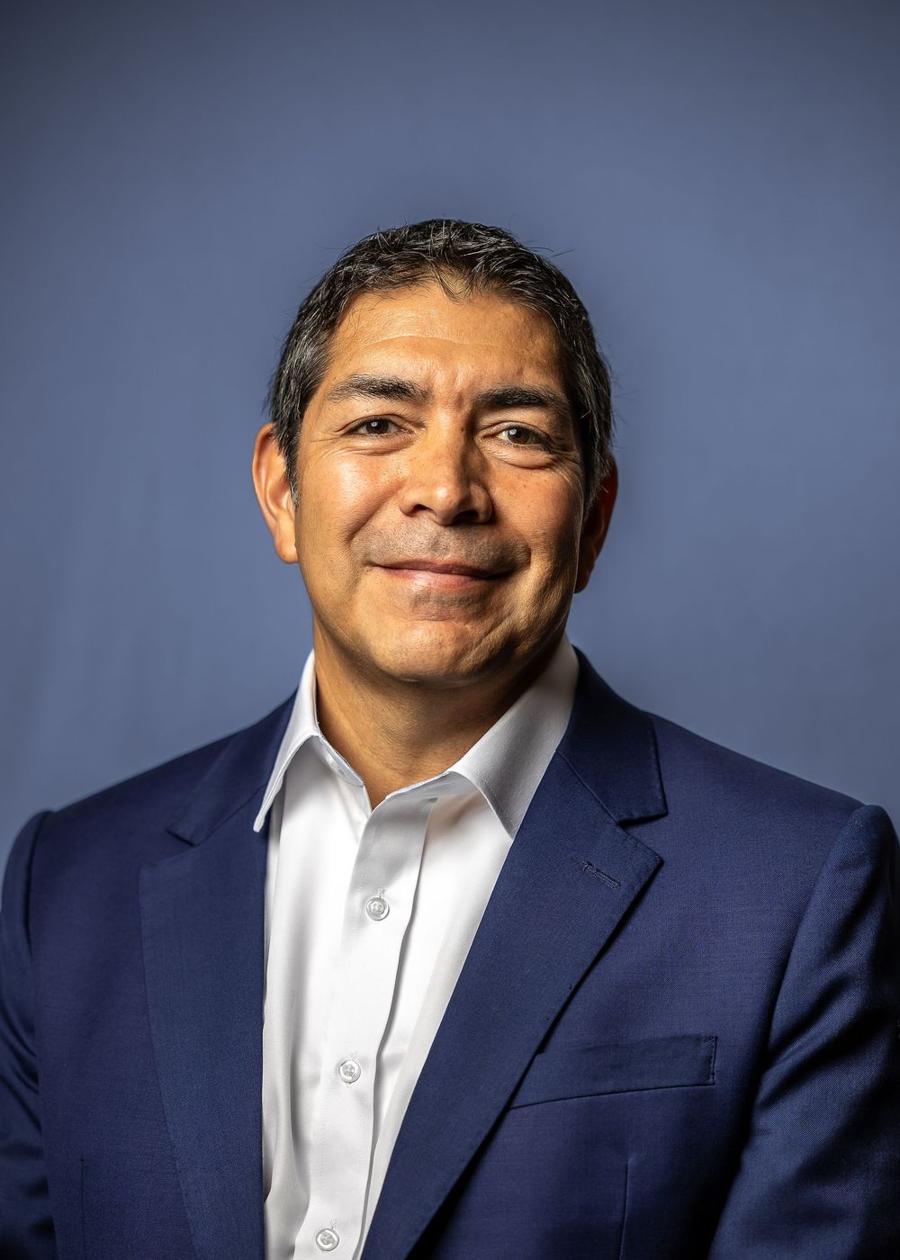 Professional headshot of someone wearing a navy suit and white shirt against a blue background.