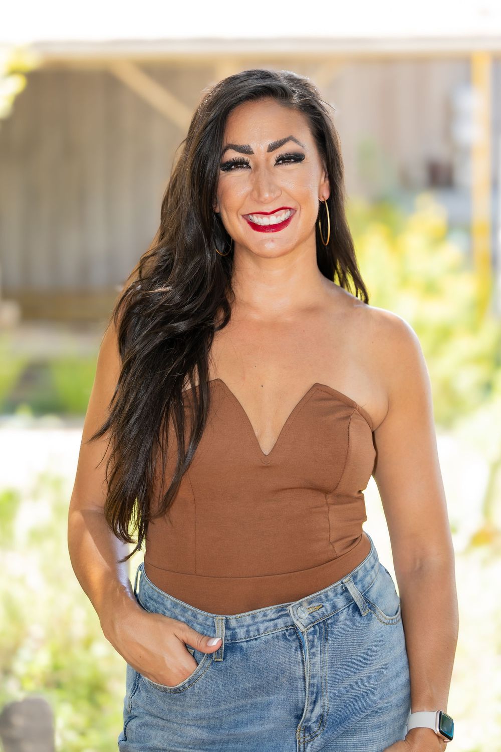 Smiling woman in brown strapless top and denim jeans poses outdoors with hand in pocket.