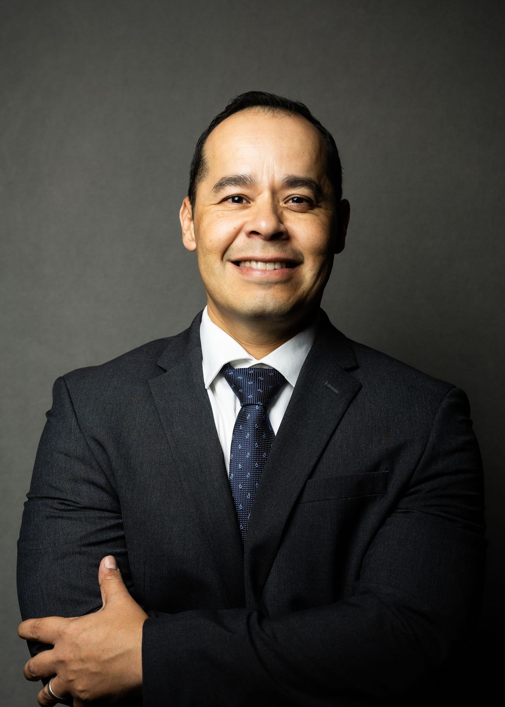 Professional headshot of a businessman in a dark suit and blue polka dot tie against a gray background.