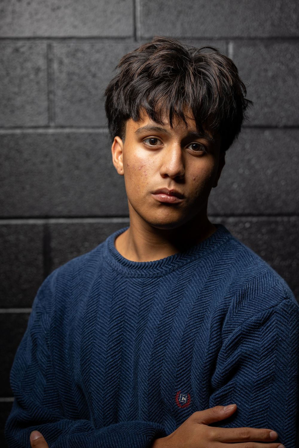 A person wearing a dark blue knit sweater poses against a gray wall in dramatic lighting.