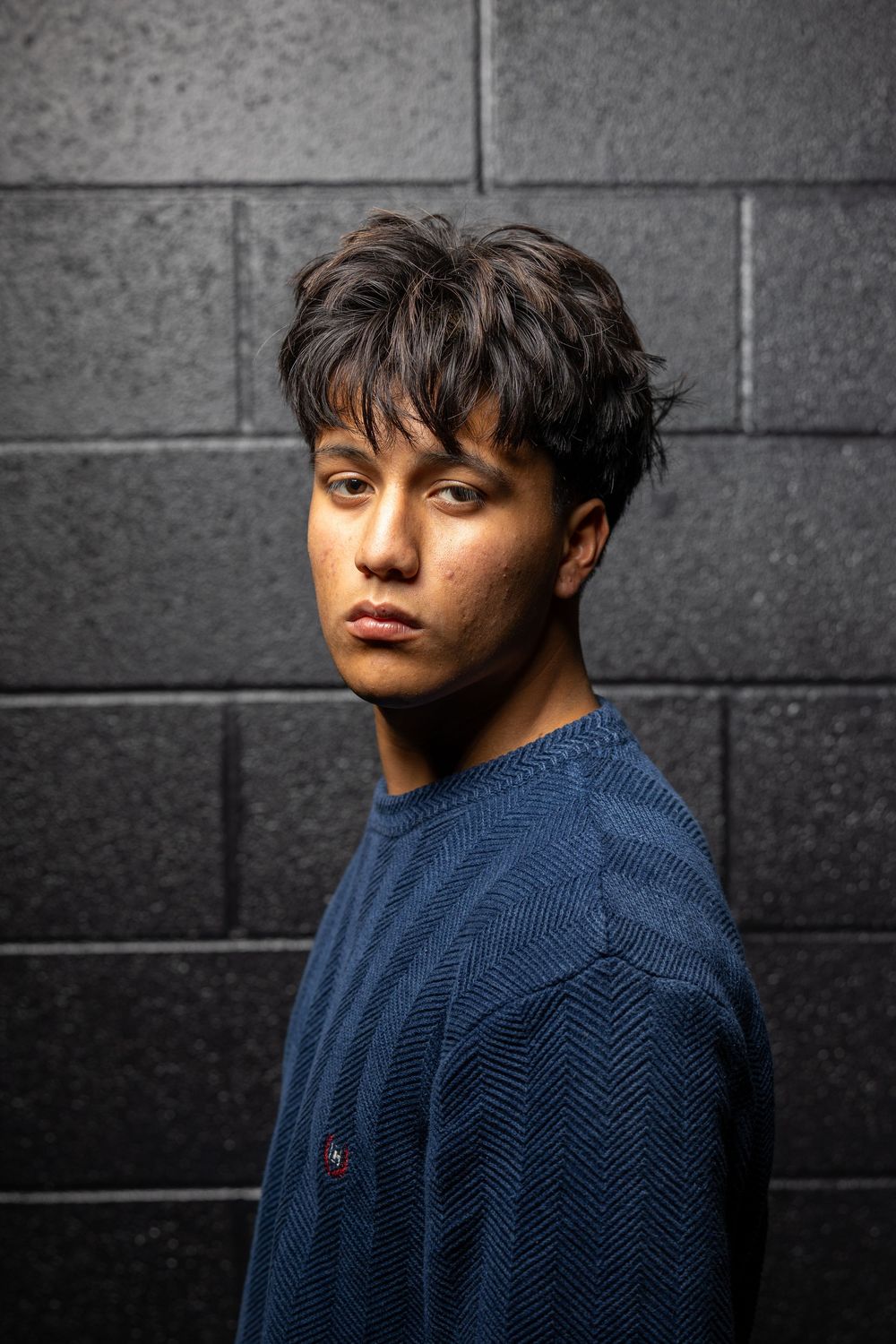 Portrait of person wearing blue sweater against dark brick wall with dramatic lighting and textured hair.