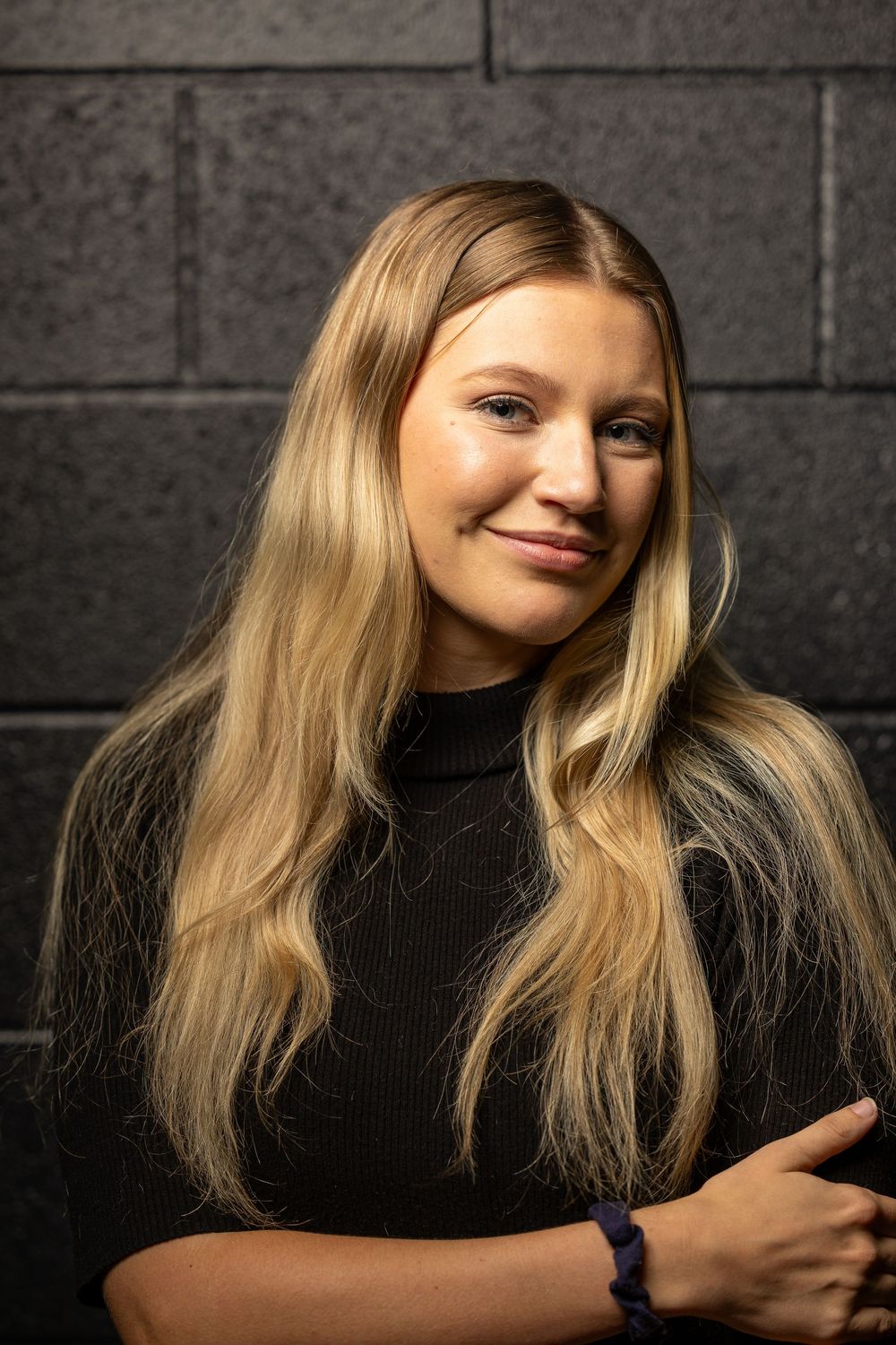 Portrait of someone with long blonde hair wearing a black sweater against a dark wall, lit by dramatic lighting.