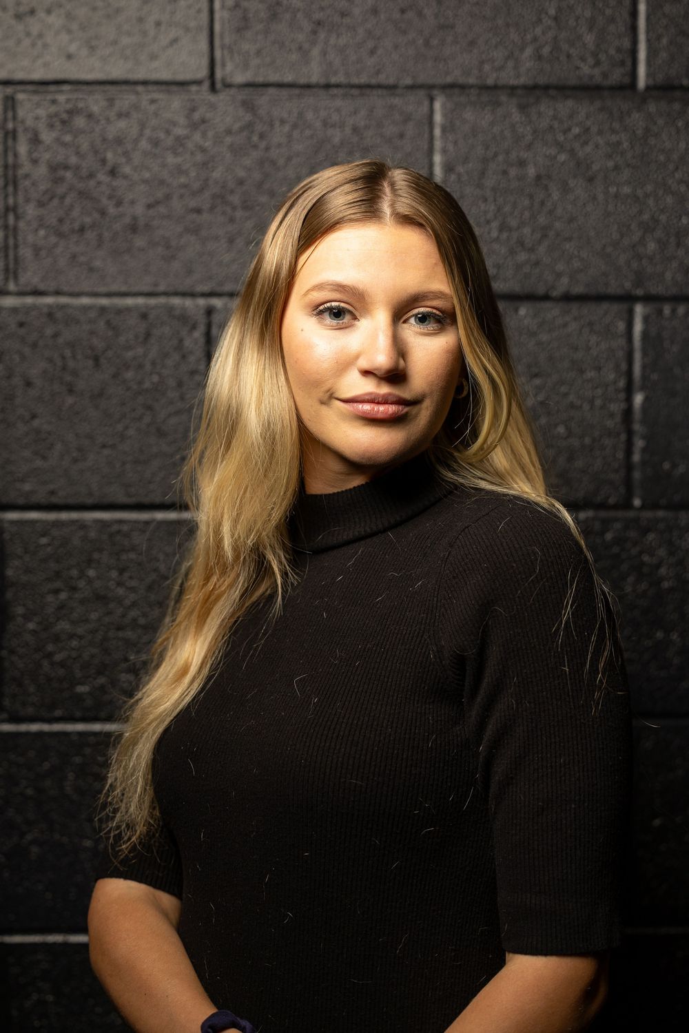 Portrait of a blonde woman in a black turtleneck sweater against a dark brick wall with dramatic lighting.