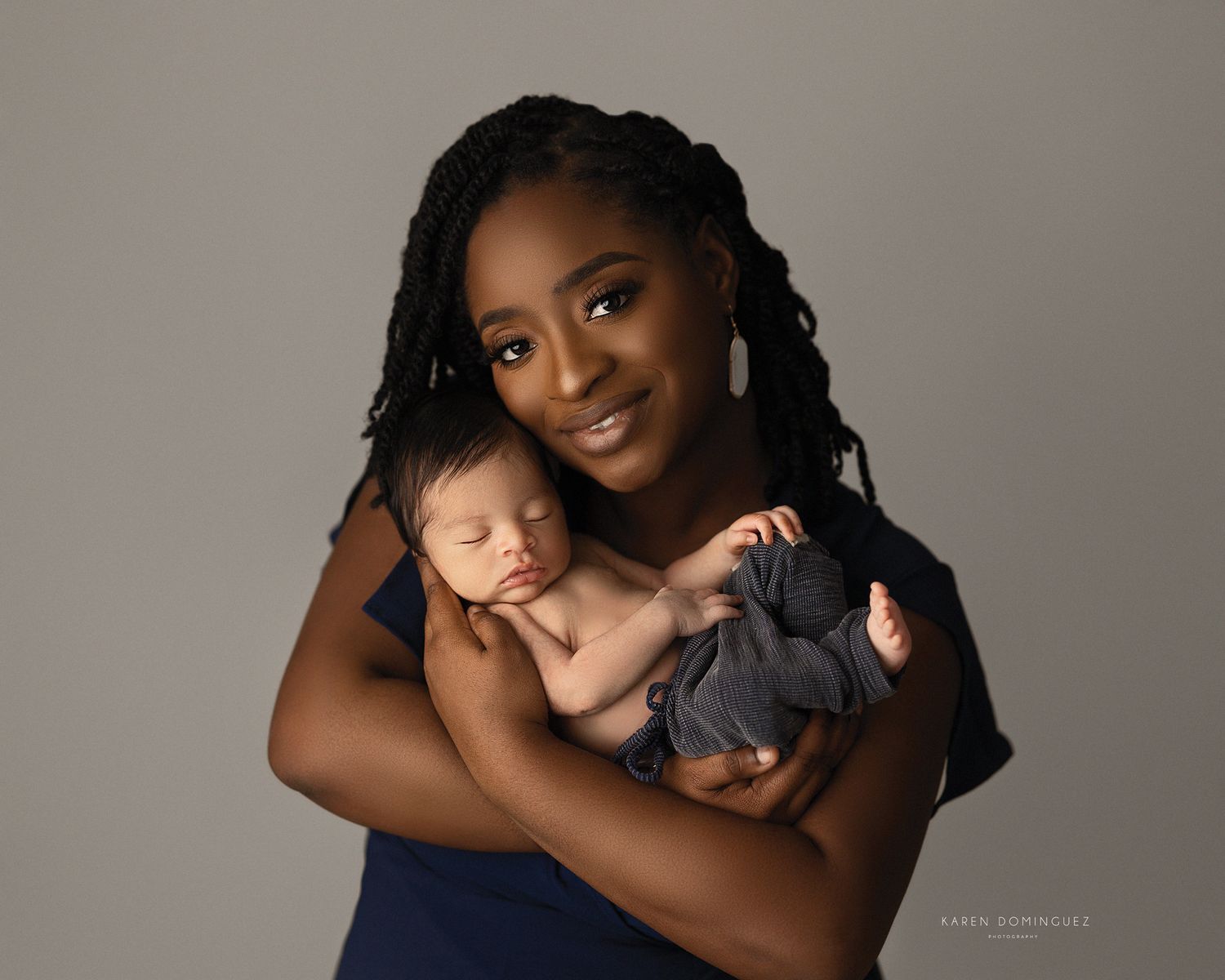 Caring mother in navy blue dress holds sleeping newborn baby during professional portrait session against gray backdrop.