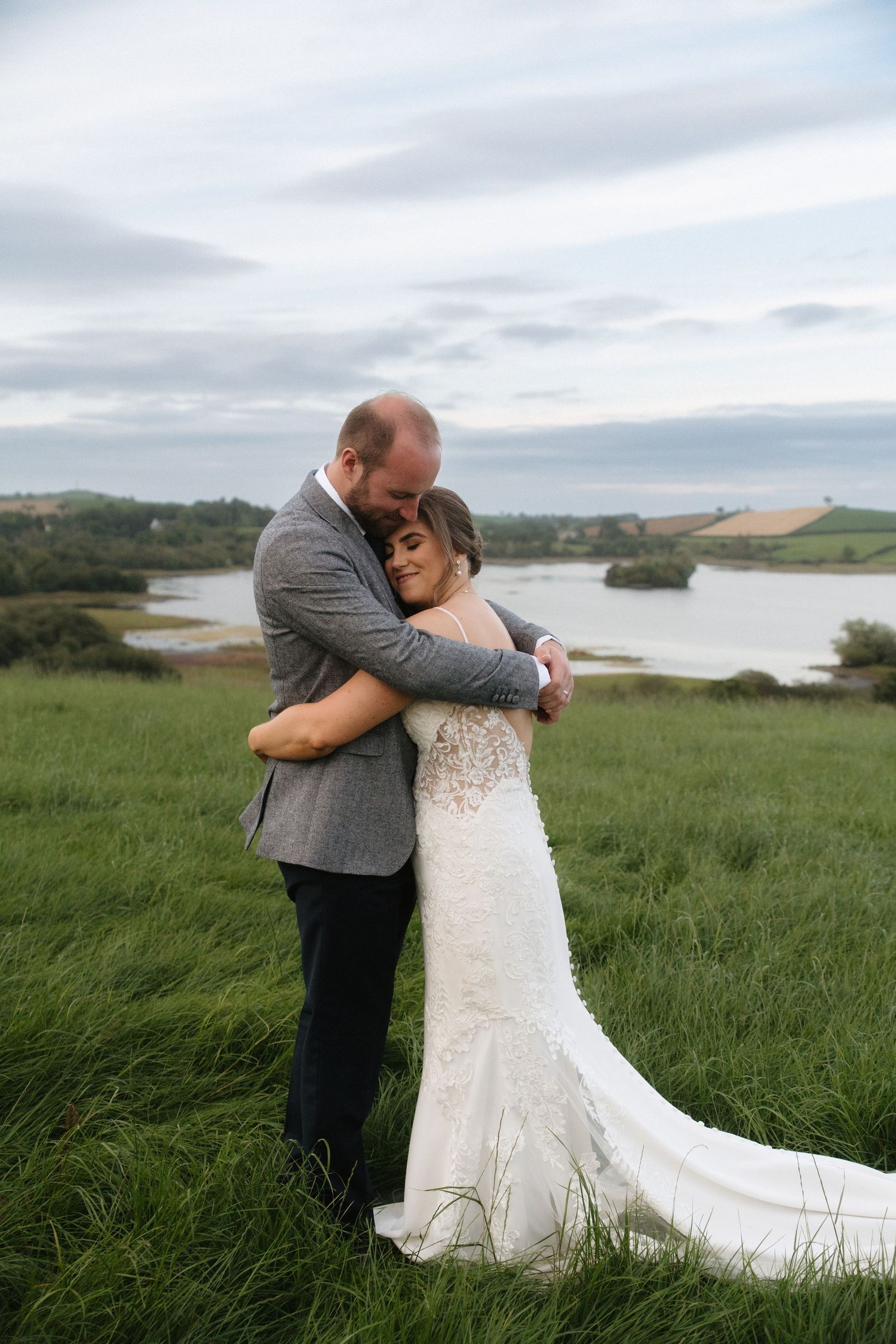 A couple embraces on a grassy coastal cliff overlooking a serene bay.