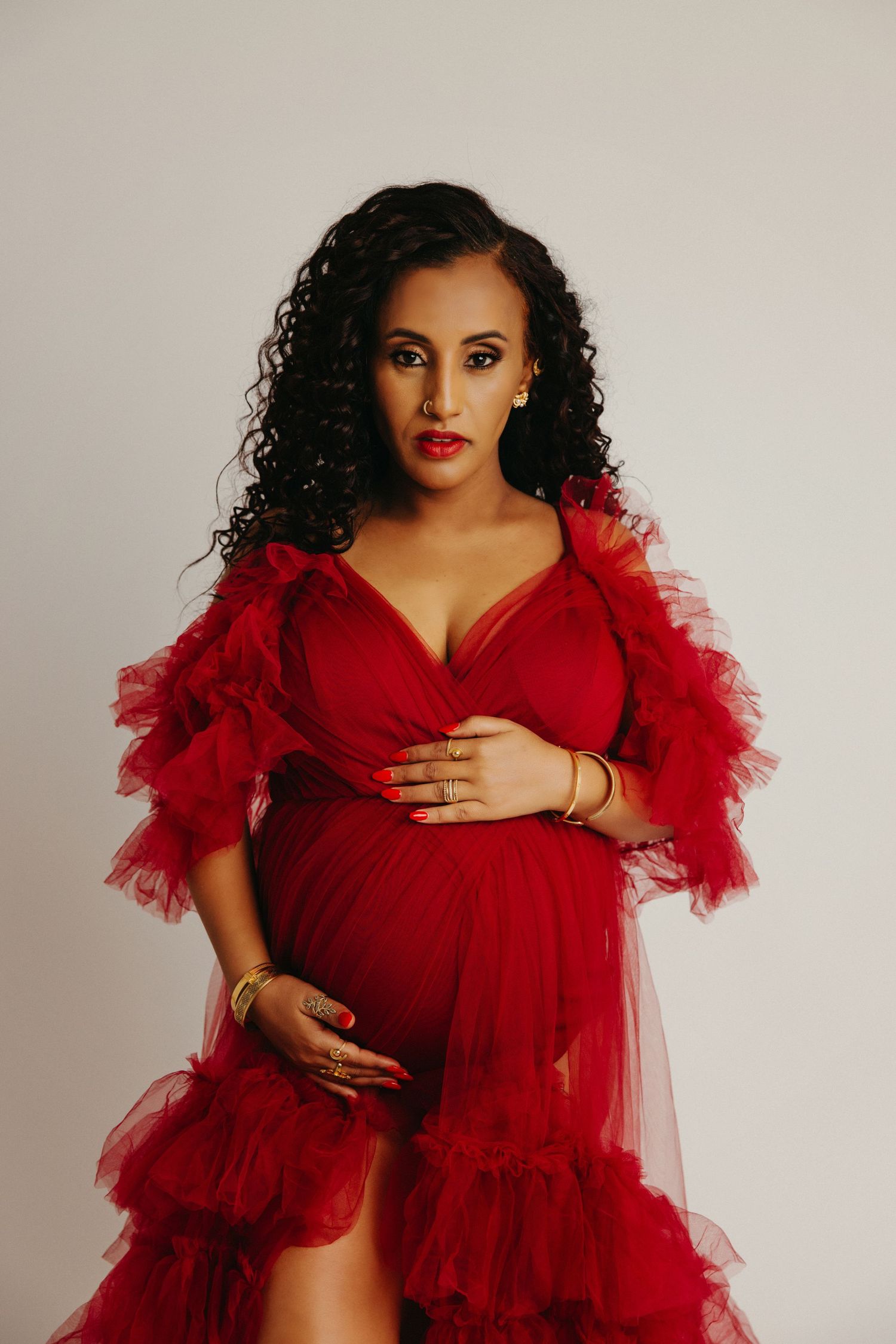 Elegant maternity portrait featuring a flowing red tulle dress with dramatic ruffled sleeves against a neutral backdrop.