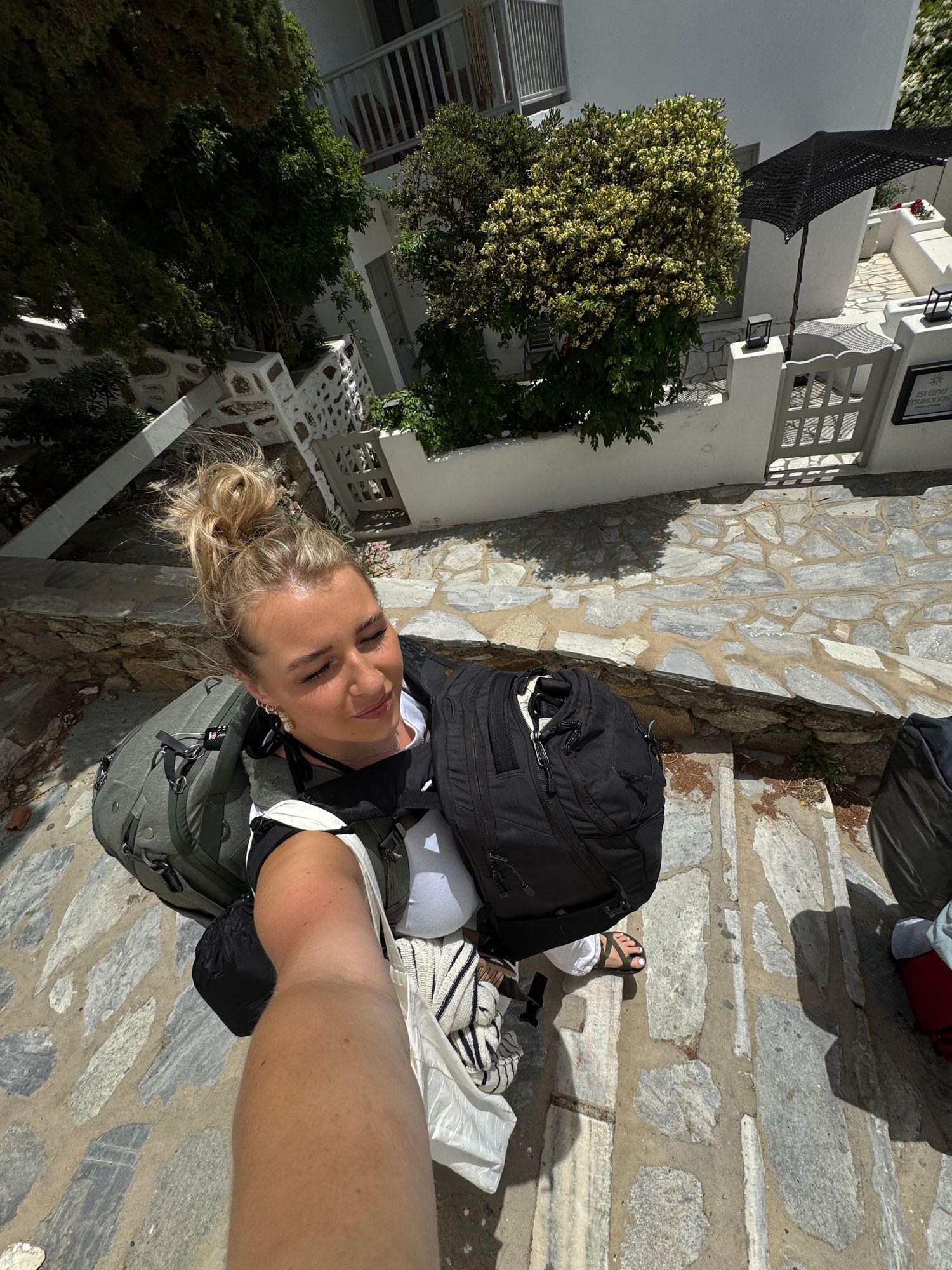 A person taking a selfie on stone steps with white buildings and greenery in background.