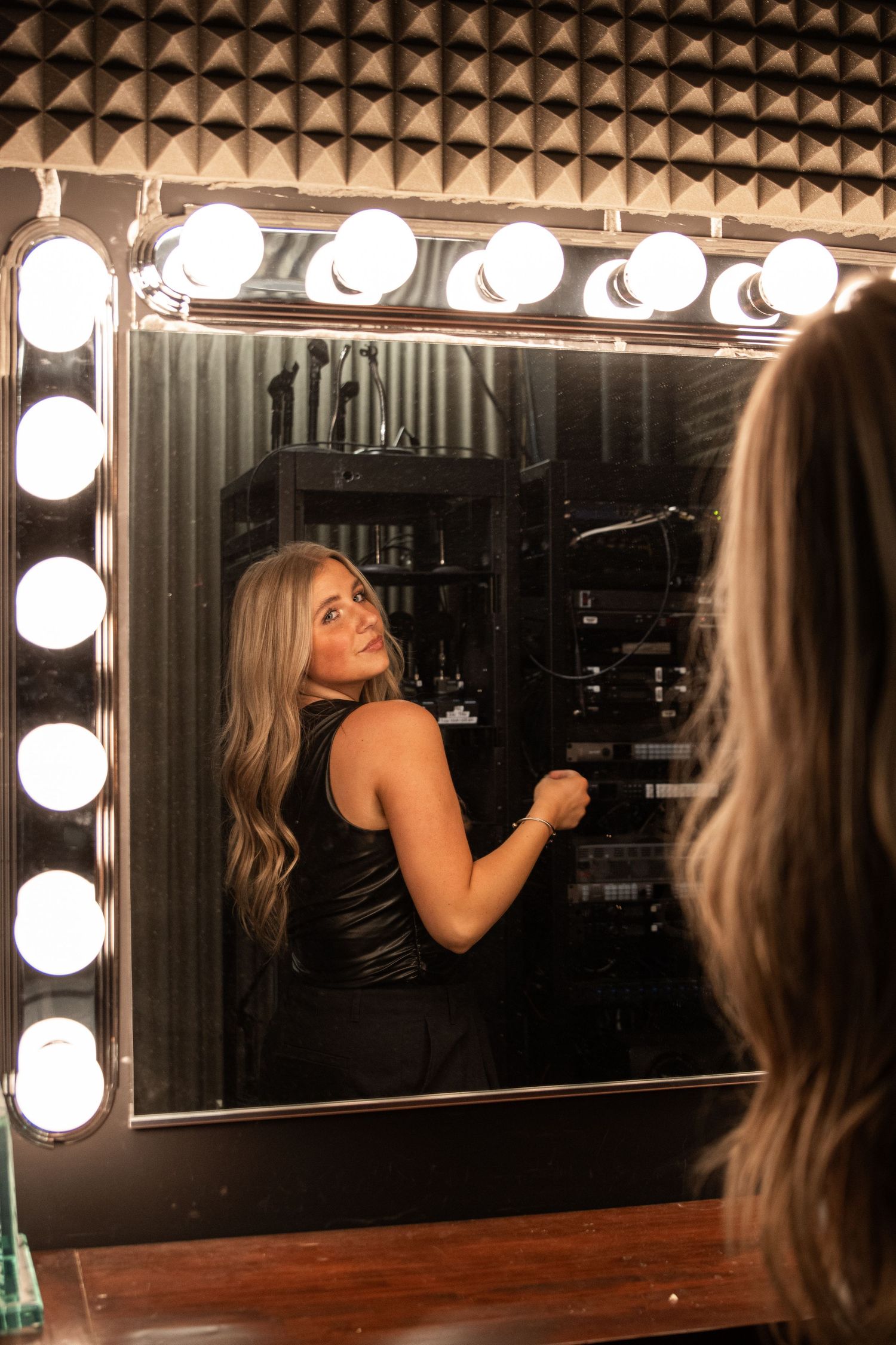 Cora Emilie Media Person looking over shoulder at reflection in lighted vanity mirror with round bulbs.