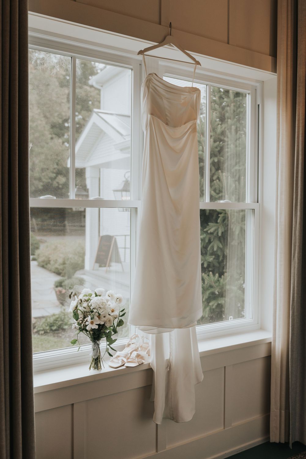 White wedding dress hanging in front of sunlit windows with soft pink roses on the windowsill.