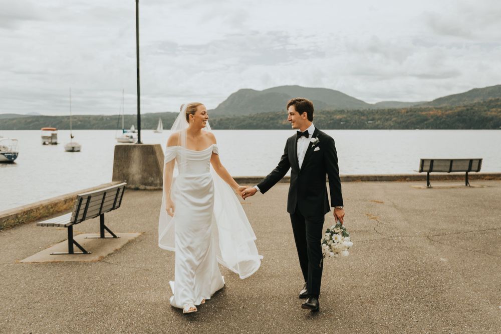 A couple in wedding attire walks along a scenic coastal path with mountains and water in the background.