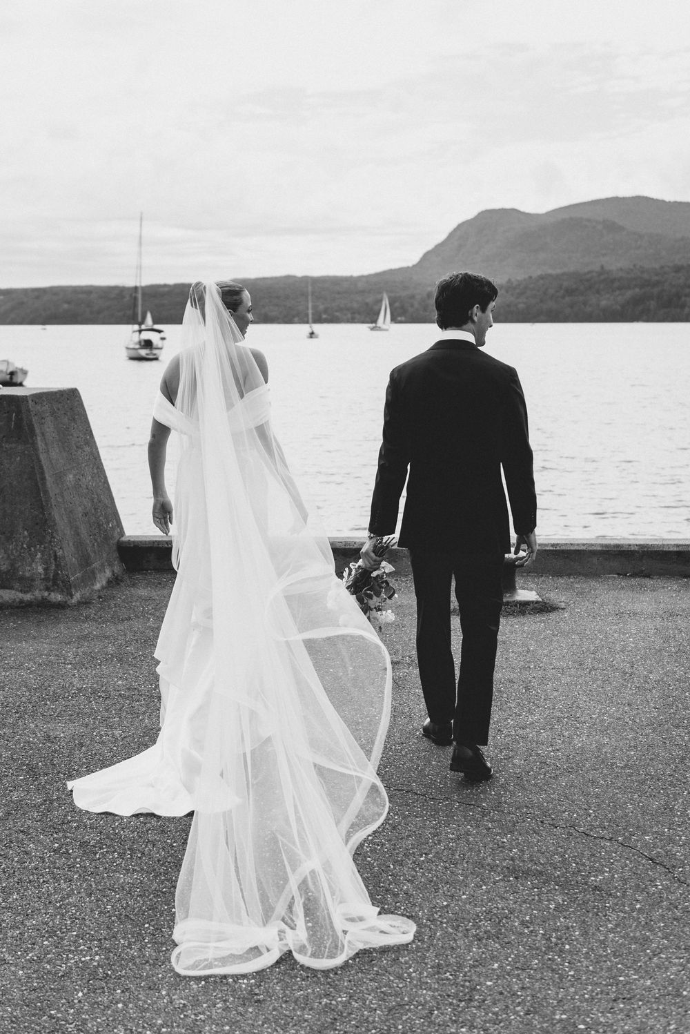 A wedding couple stands on a pier overlooking a lake with sailboats and mountains in the background.