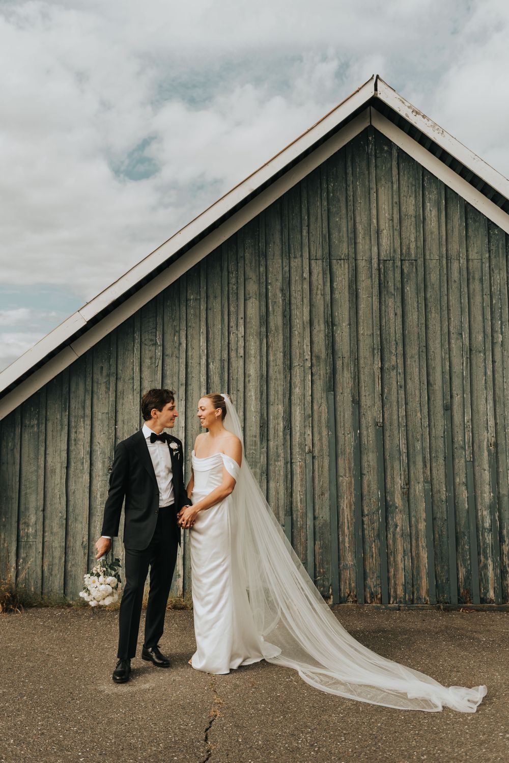 Newlyweds pose in front of a rustic wooden barn with a flowing white veil trailing behind the bride.