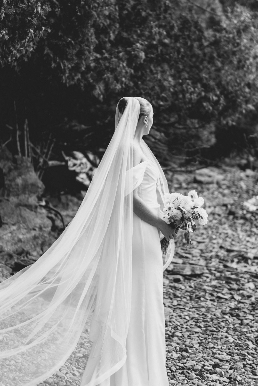 A bride in a flowing white wedding dress and long veil stands in a garden setting, holding a delicate floral bouquet.