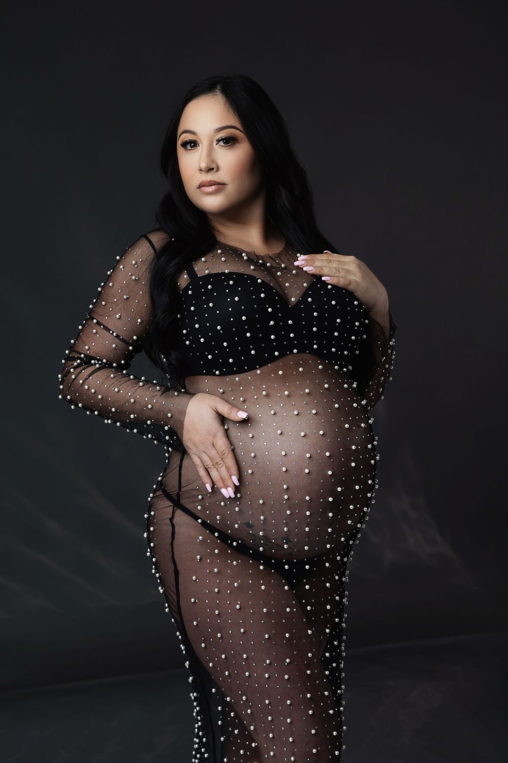 Maternity portrait in sheer black dotted gown against dark background.