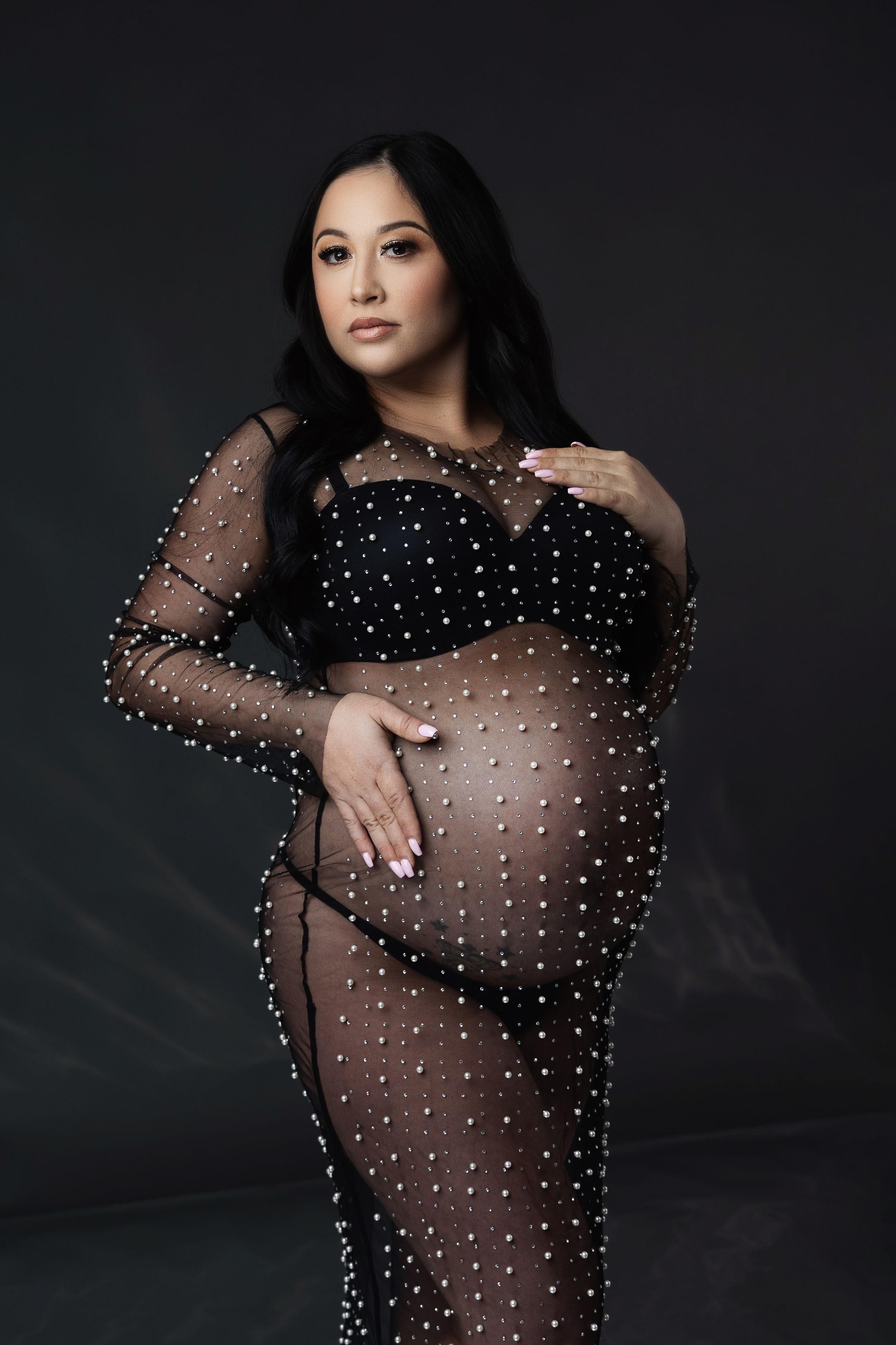Maternity portrait in sheer black dotted gown against dark background.