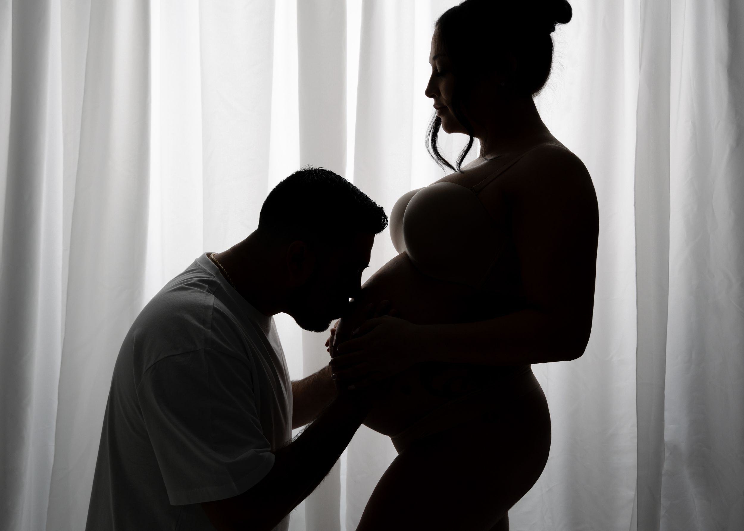 Silhouette of an intimate maternity moment between two people against a backlit curtained window.