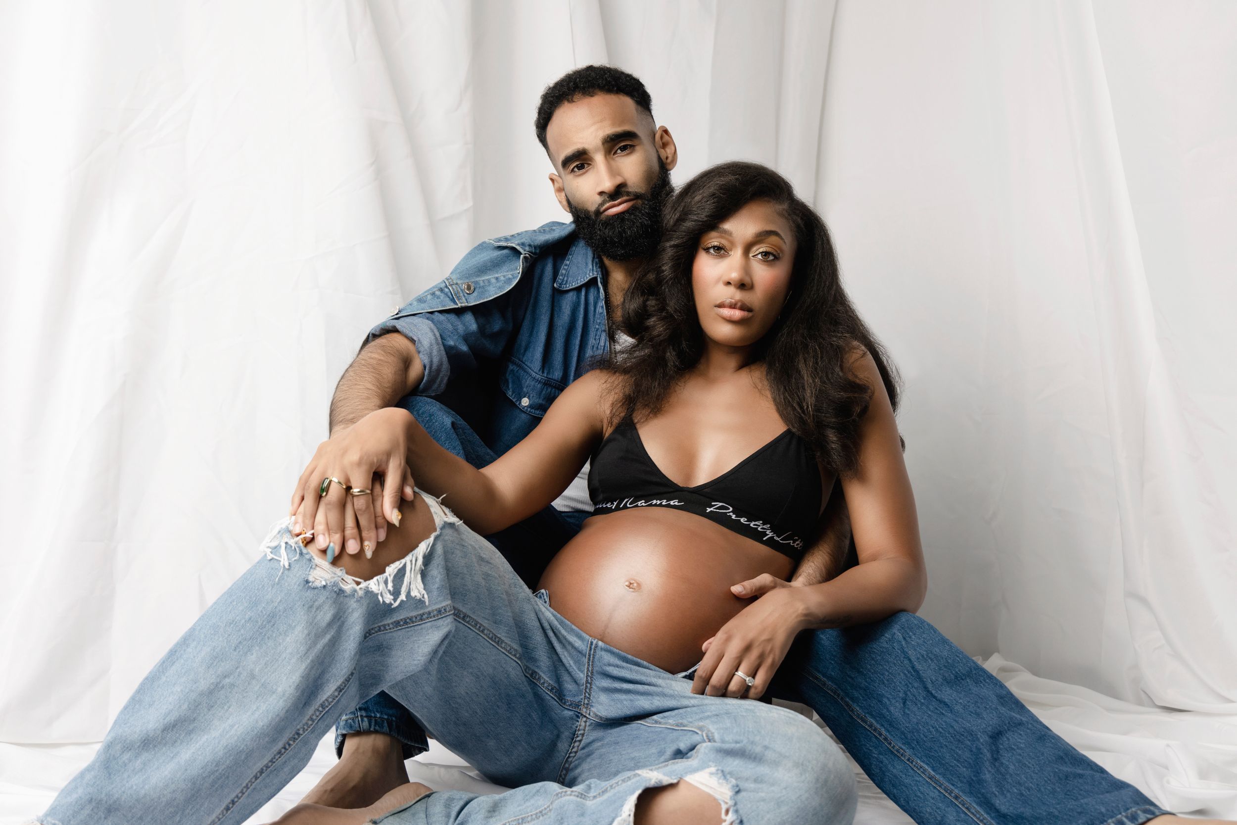Intimate maternity portrait of couple in casual denim against white backdrop.