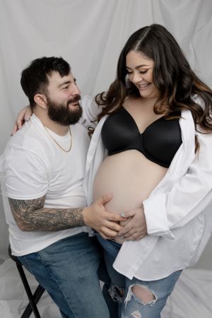 A loving maternity photo of a couple in white shirts against a light backdrop.