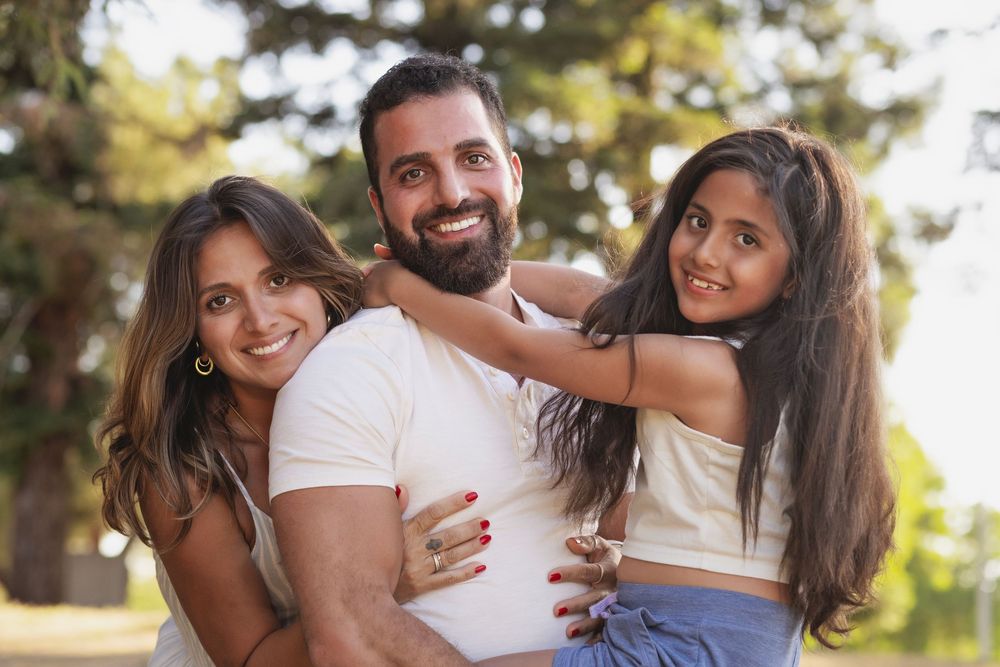 Happy family of three smiling outdoors in a park, parents holding young daughter between them.