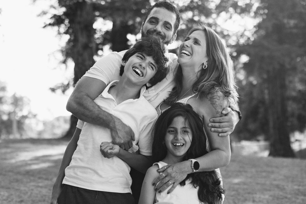 Black and white photo of a joyful family of four hugging and laughing together in a park.