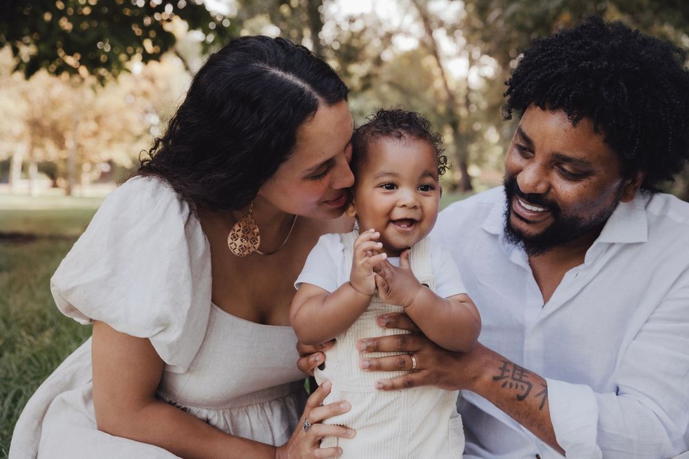 Happy family with mother kissing baby girl while father smiles in a sunny park setting.