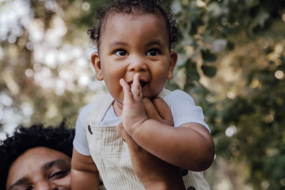 Adorable baby with hands near mouth being held by parent outdoors among green trees.