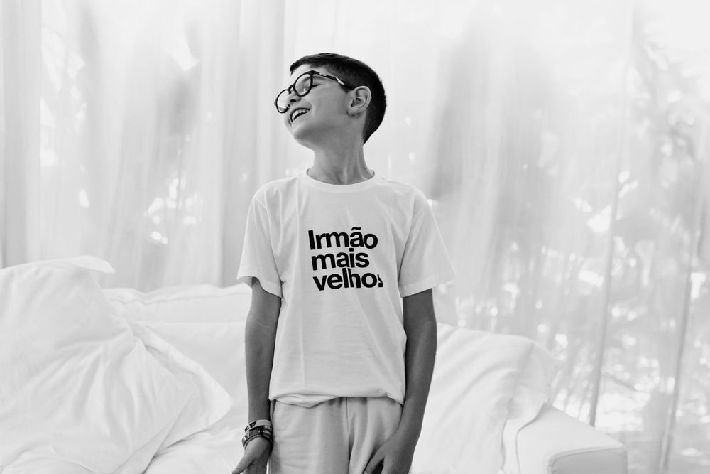 Young man wearing glasses and 'Irmão mais velho' t-shirt smiling in black and white photo.