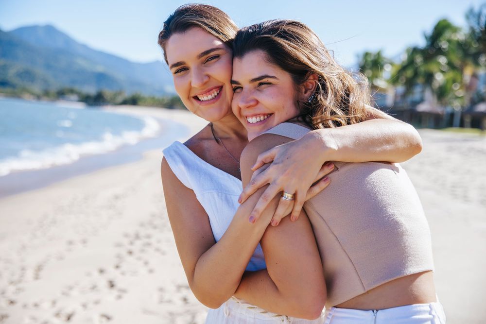Two women hugging and smiling on a sunny beach with mountains and palm trees in the background.