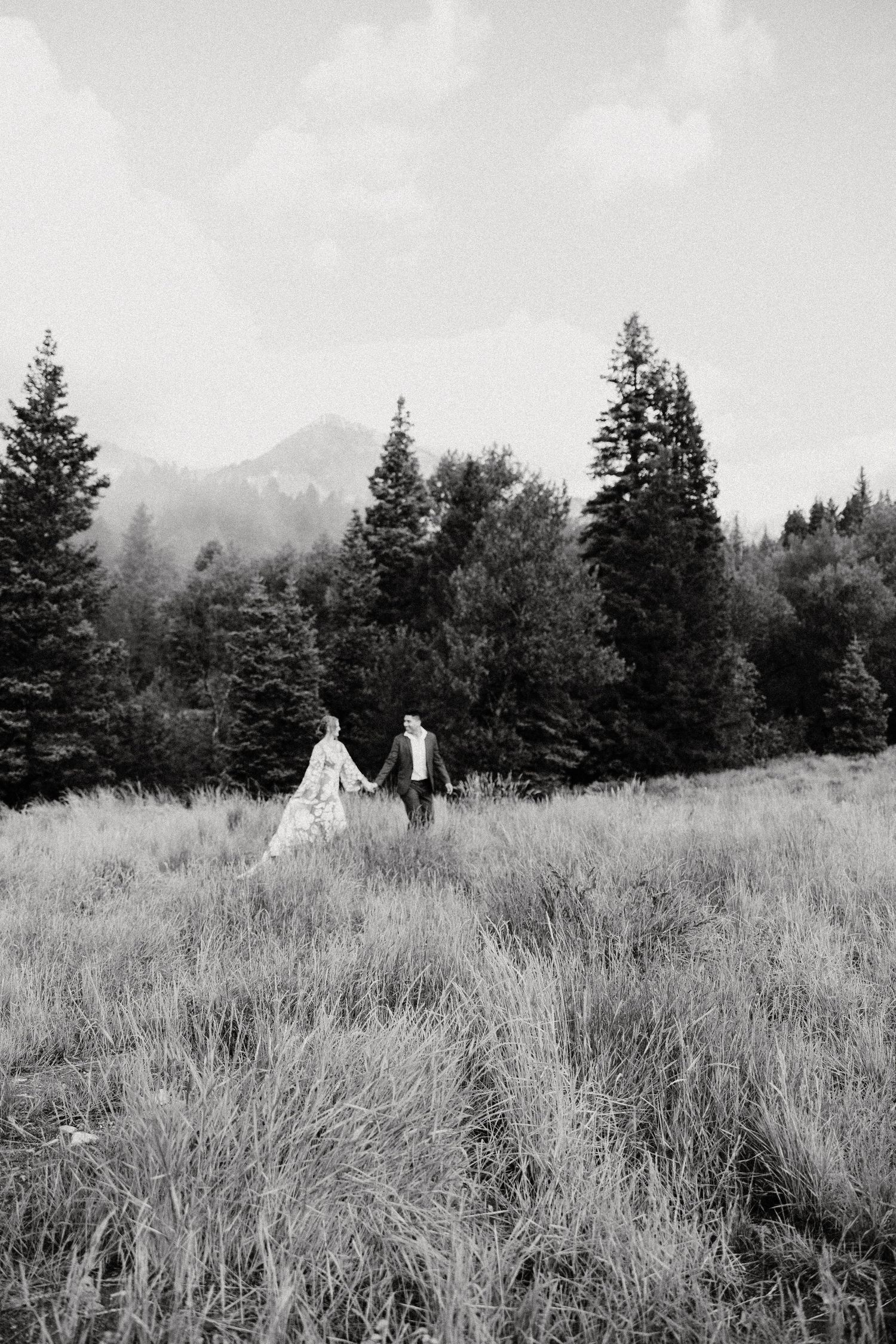 Black and white photo of a couple walking through mountain meadow surrounded by evergreen trees. Elopement, Mountains,