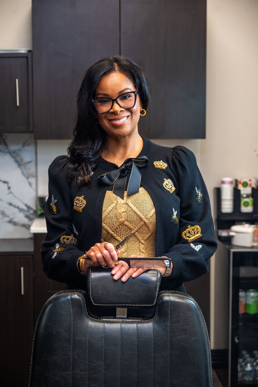 A hairstylist places her hands on top of her styling chair while smiling for a portrait inside her hair salon.
