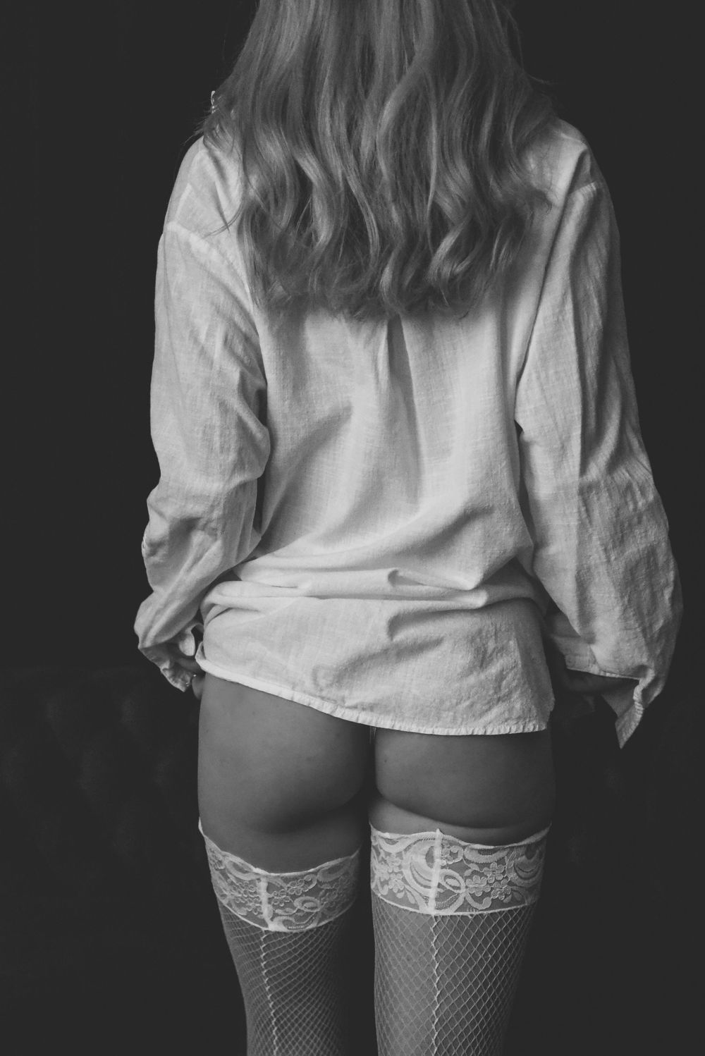 Black and white photo showing a person from behind wearing a white button-down shirt and lace stockings.