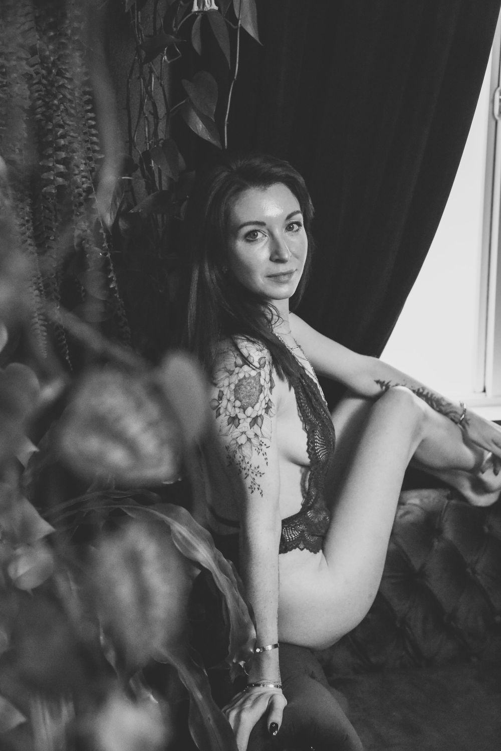 Black and white boudoir photo featuring lace lingerie and dramatic lighting against dark curtains.