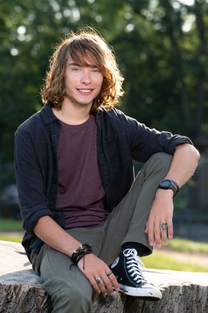 A casual portrait of a teenager sitting on a tree stump wearing a black cardigan and Converse sneakers.