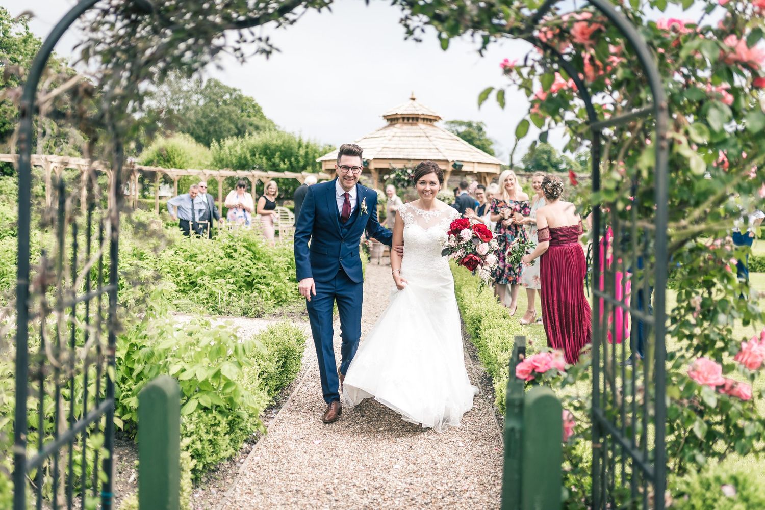 A couple walks through a garden archway lined with blooming flowers on their wedding day.