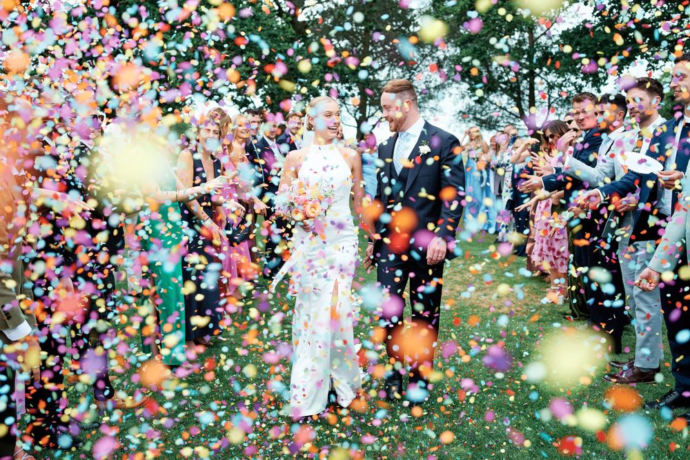 Bride and groom walk through colorful confetti shower surrounded by wedding guests outdoors.