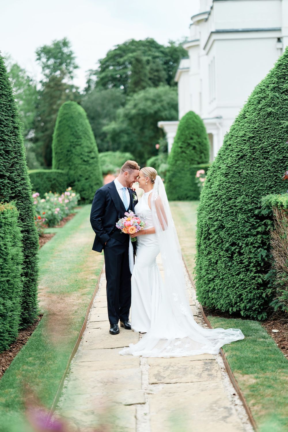 Bride and groom share a romantic moment on a garden path lined with manicured hedges, bride holding colorful bouquet.