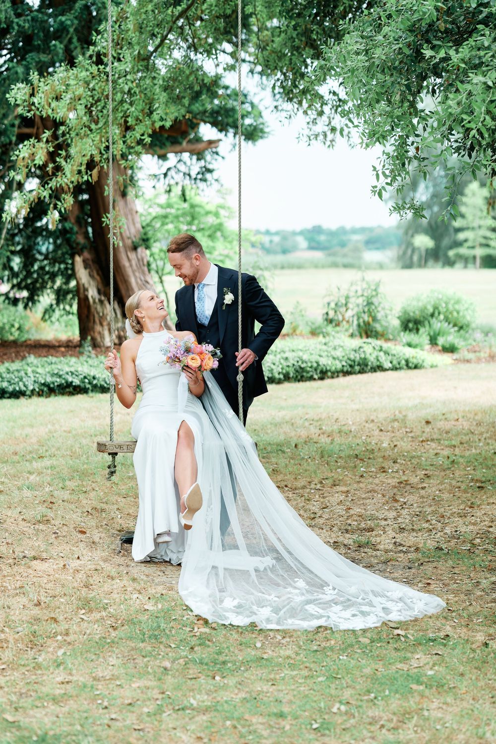 Bride in white gown sits on tree swing as groom in navy suit leans in close, surrounded by lush greenery.