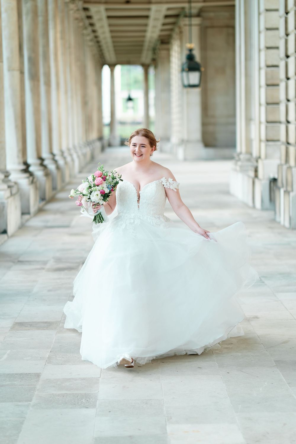 Smiling bride in off-shoulder white ballgown holding pink bouquet walks through elegant stone colonnade.