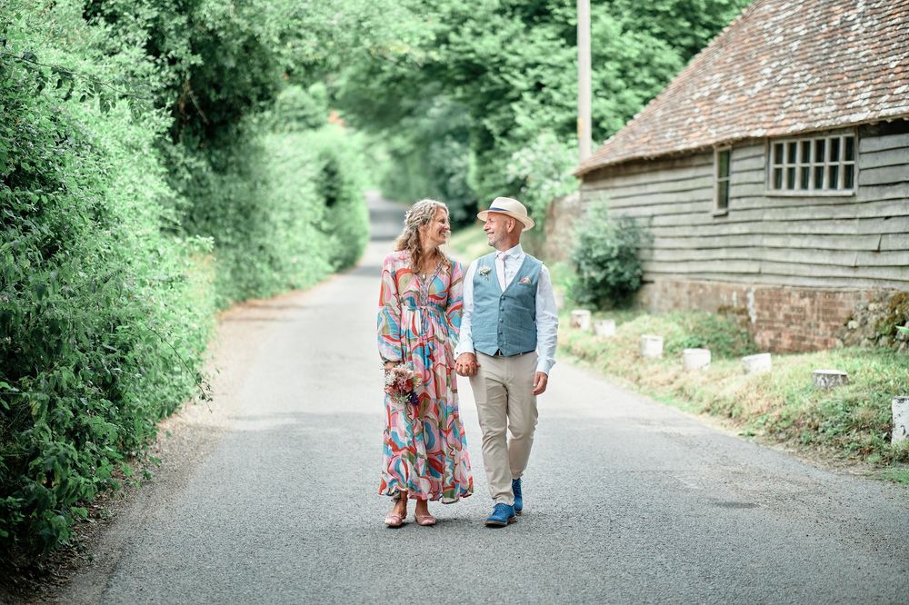 A couple walks hand in hand down a scenic countryside path surrounded by lush greenery and rustic buildings.