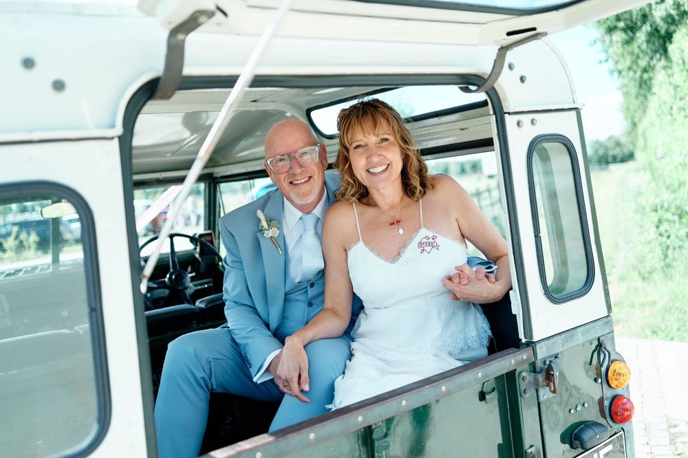 Happy couple in wedding attire smiling inside a vintage Land Rover on their wedding day.