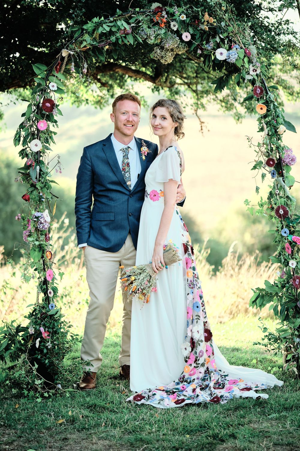A romantic wedding couple poses under a floral garden arch in an outdoor summer setting.