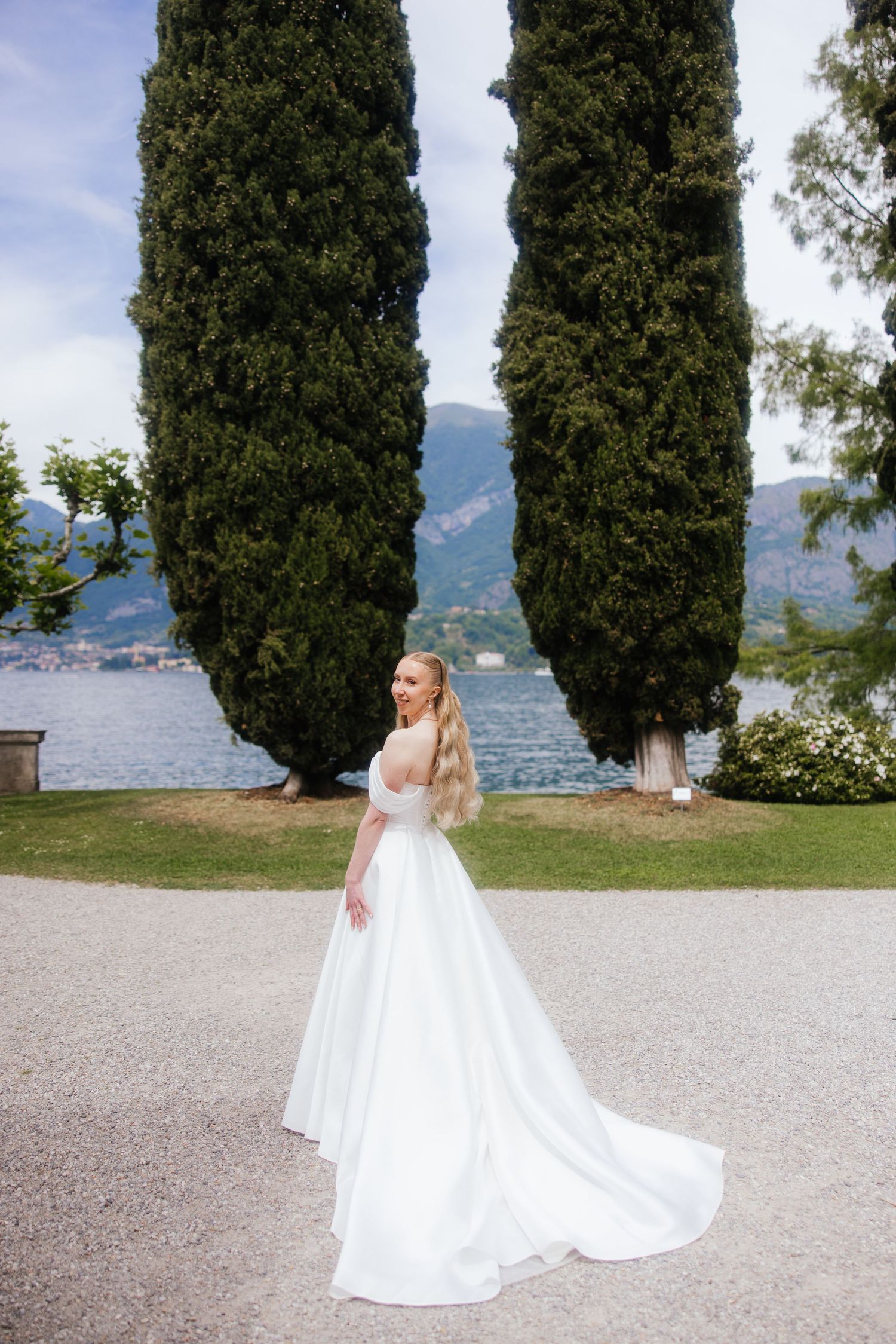 A couple in wedding attire pose together by Lake Como with Villa Balbianello in the background, Italy.