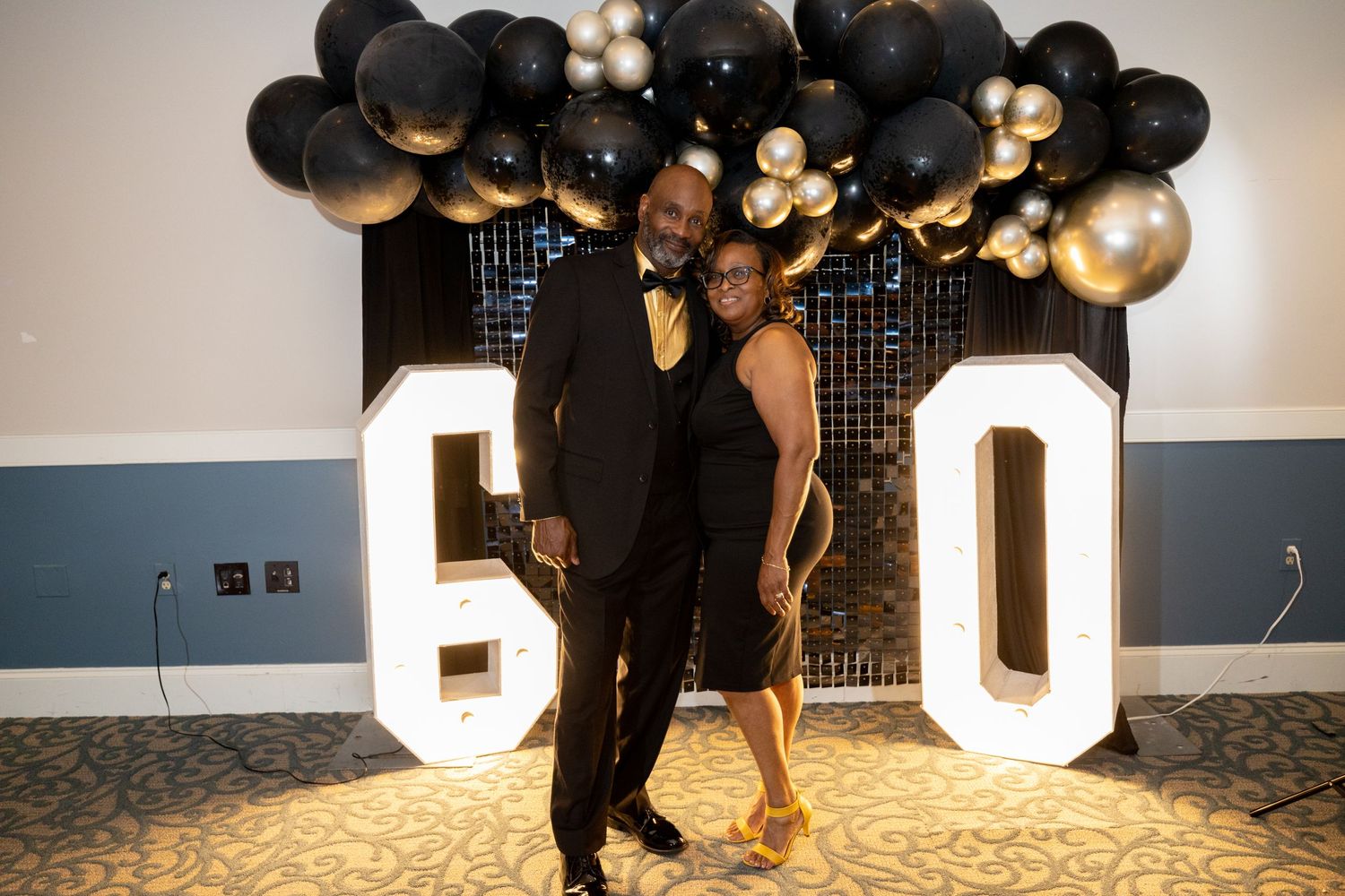 A couple poses in front of large illuminated '60' numbers with black and gold balloon decorations.