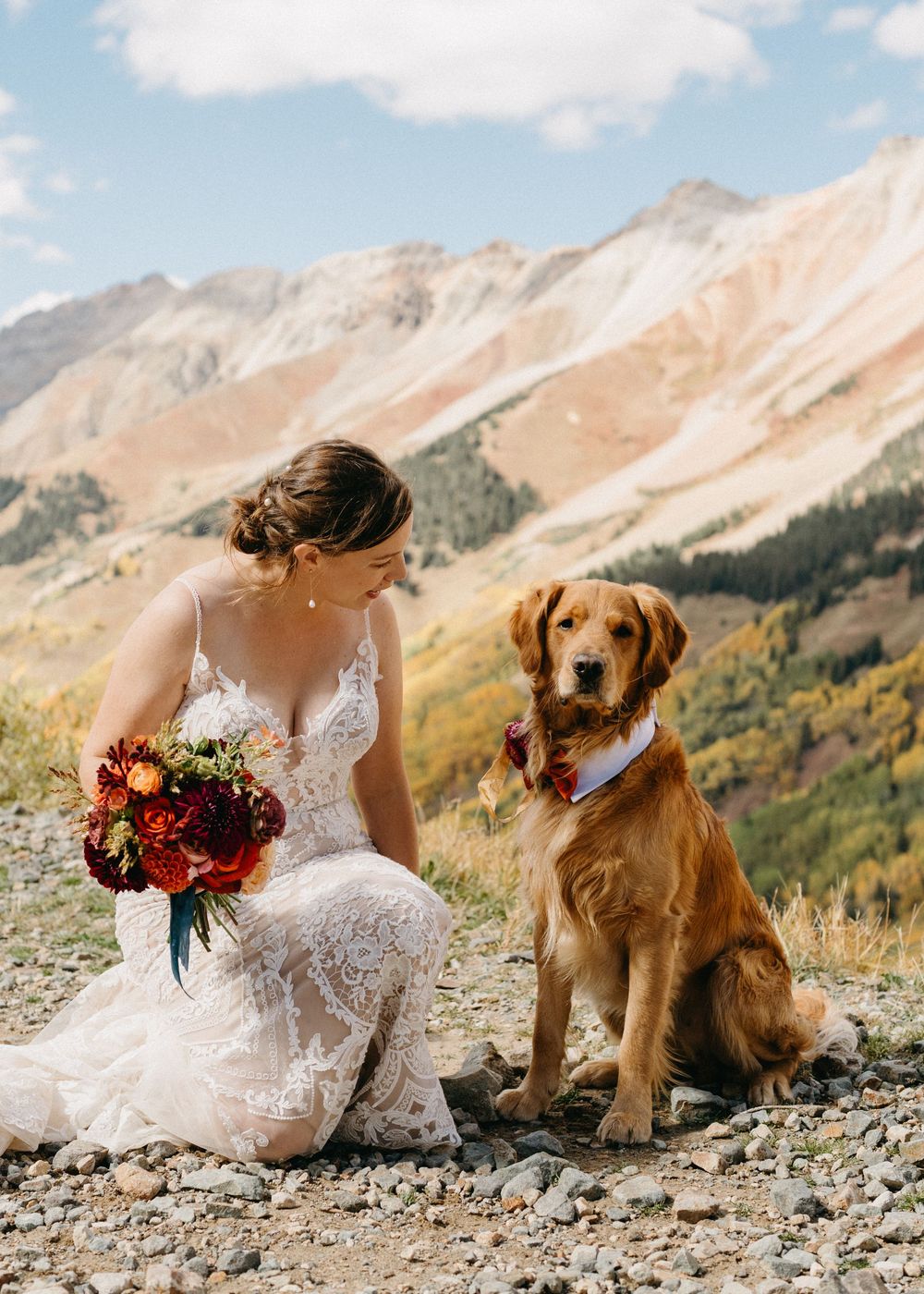 A golden retriever sits beside someone in a white dress against a backdrop of majestic mountain peaks.
