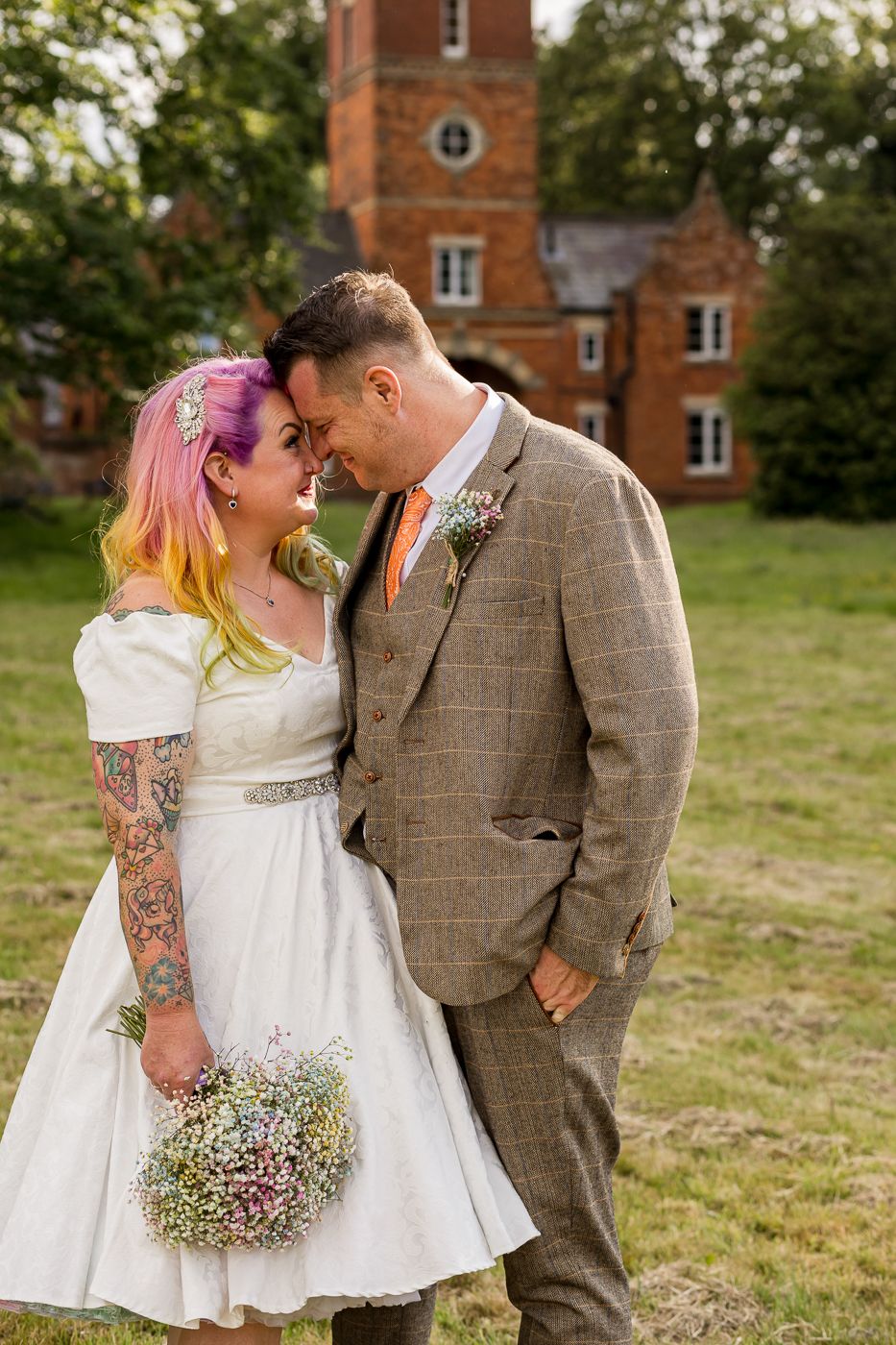Couple in vintage-inspired wedding attire embracing on grassy lawn with brick building backdrop.