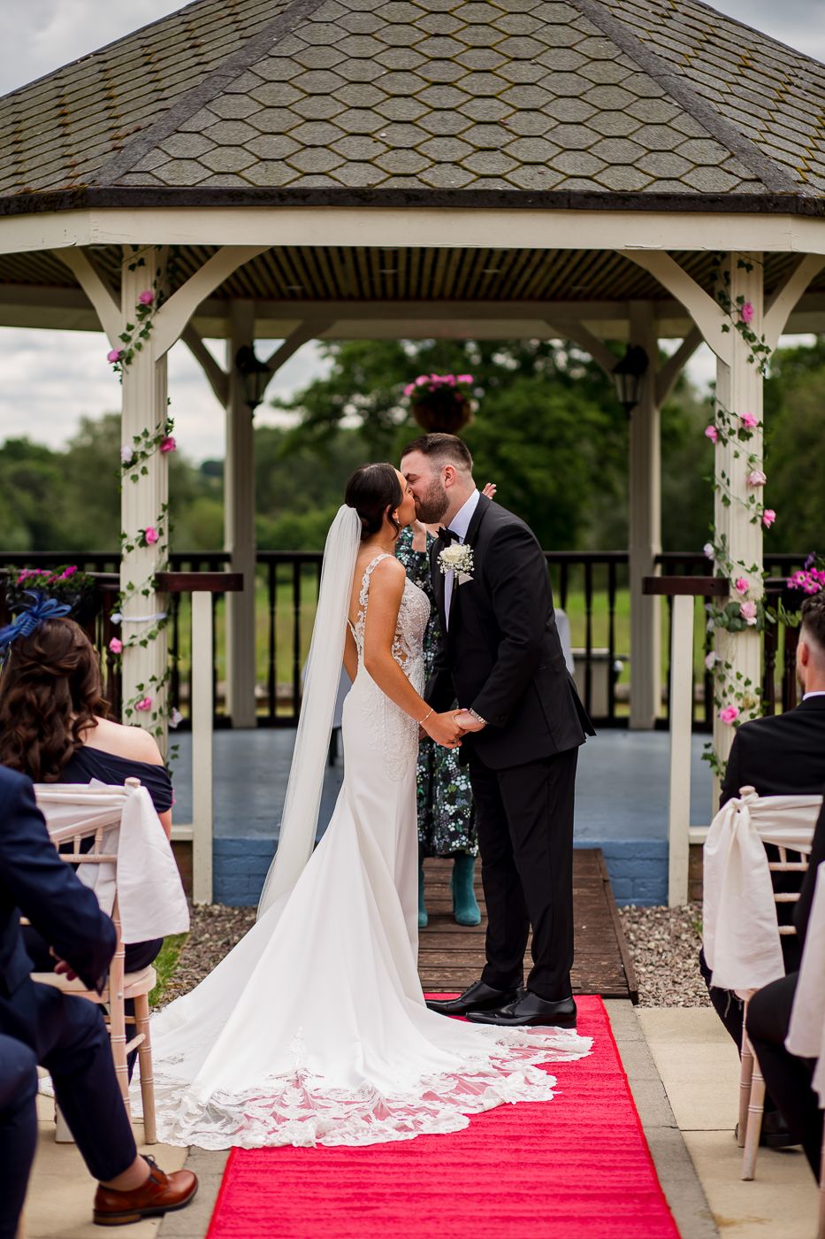 Wedding ceremony taking place under an outdoor gazebo decorated with flowers and a red carpet aisle.