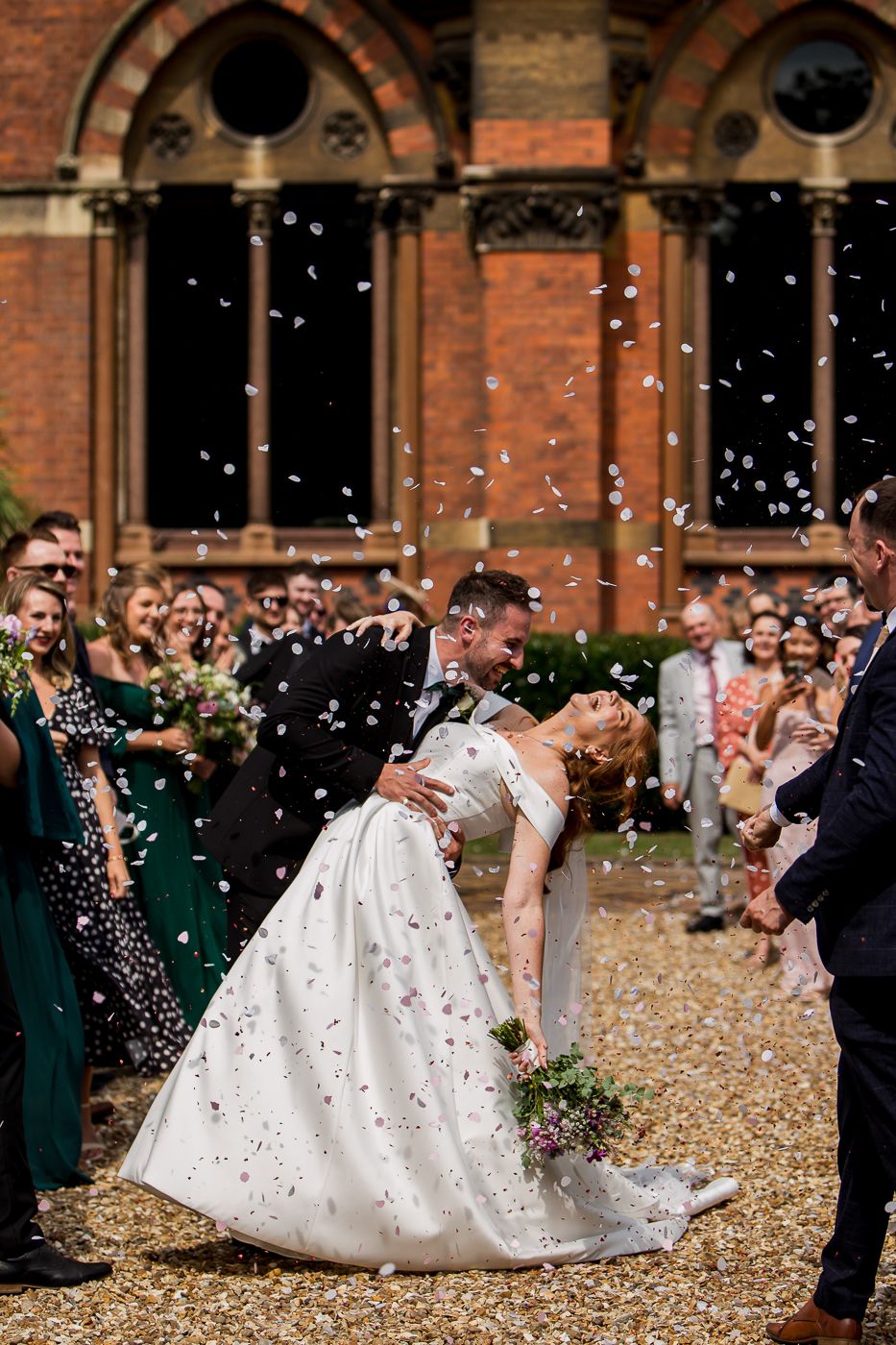 Wedding celebration at historic brick building with guests throwing confetti in joyous scene.