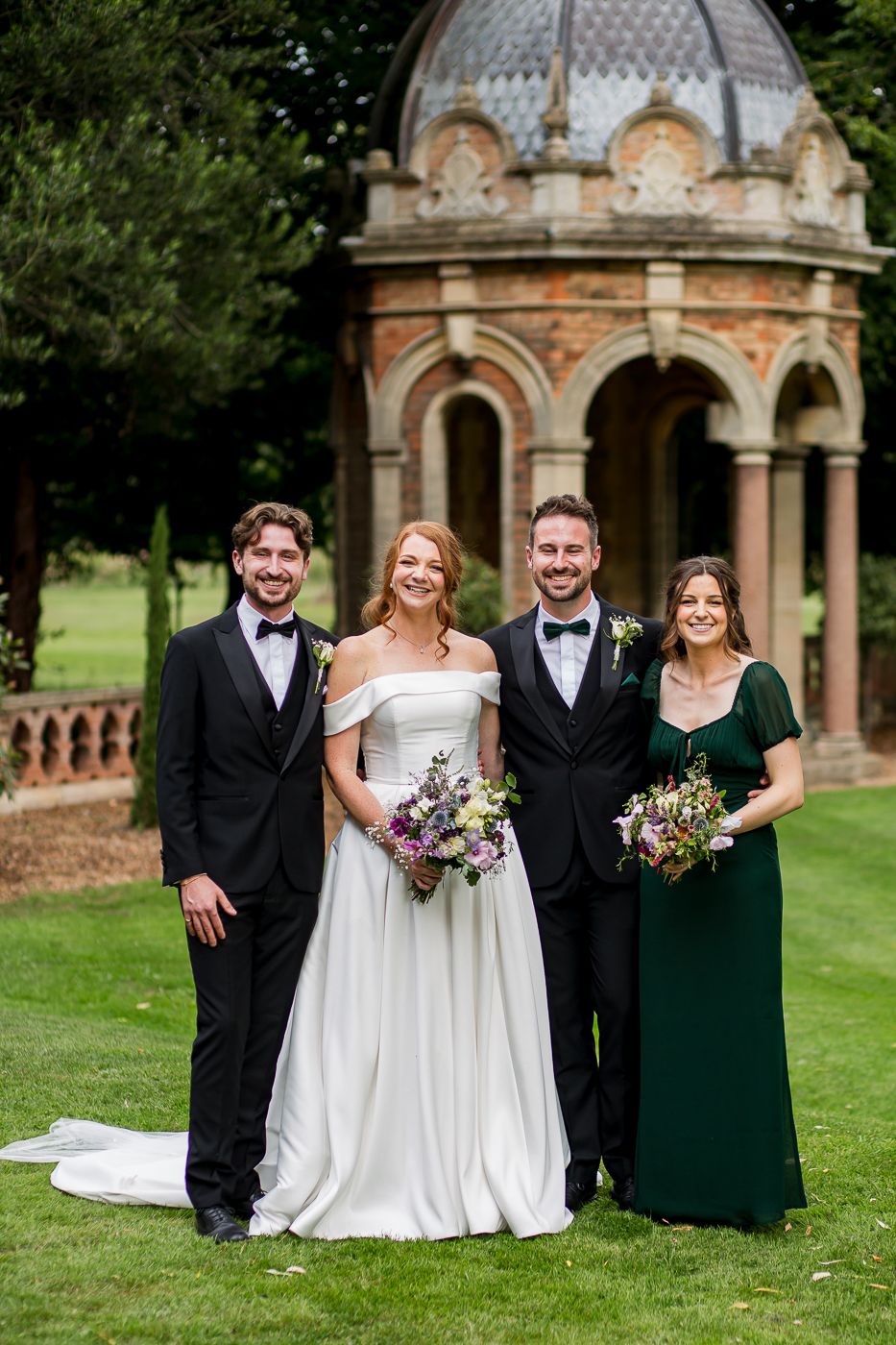 Wedding group photo in front of ornate stone gazebo surrounded by lush gardens.