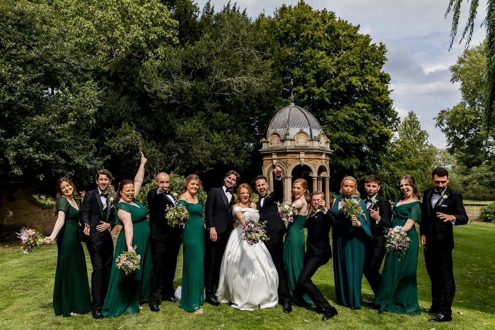 Wedding party in emerald green and black attire posed in formal garden setting.