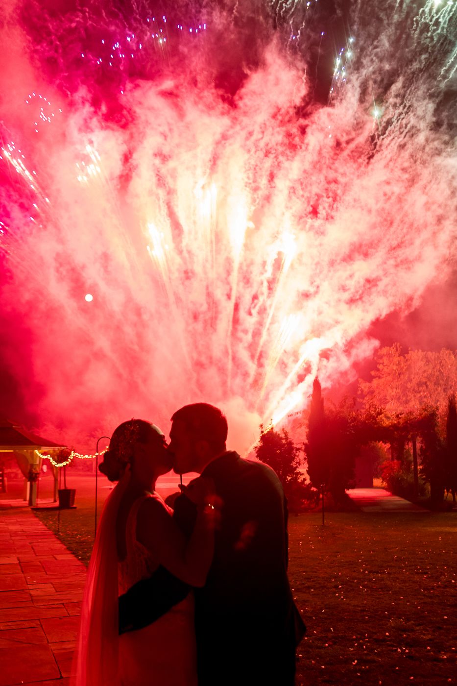 Dramatic red fireworks display silhouetting wedding celebration figures against night sky.